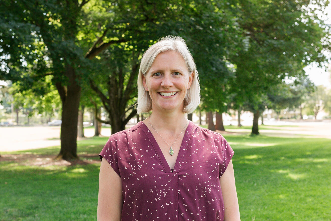 A woman with blonde hair wearing a light pink shirt smiles for her picture was standing in a park.