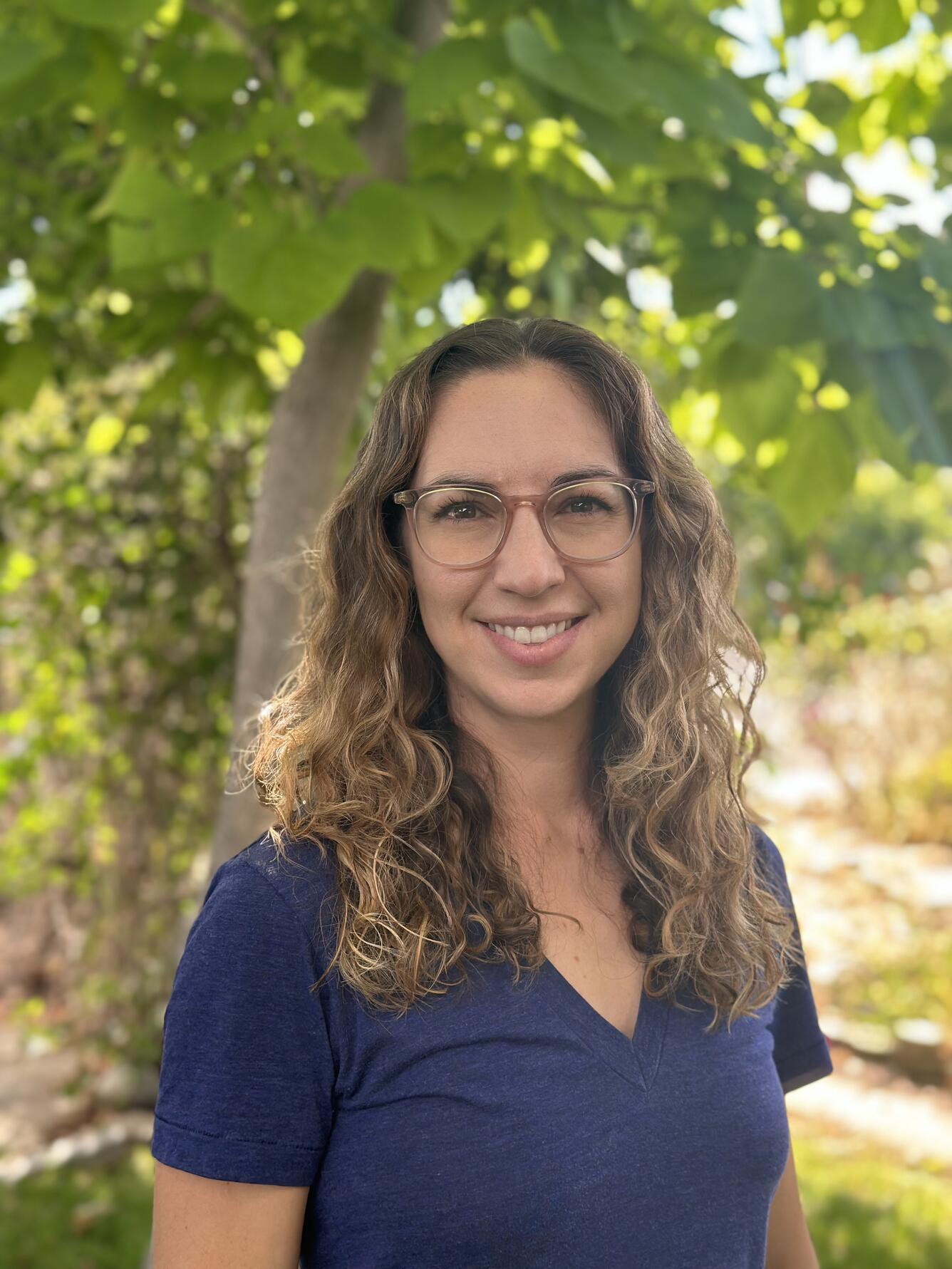 Monica is wearing a blue shirt looking at the camera. She is standing infront of a tree and smiling.
