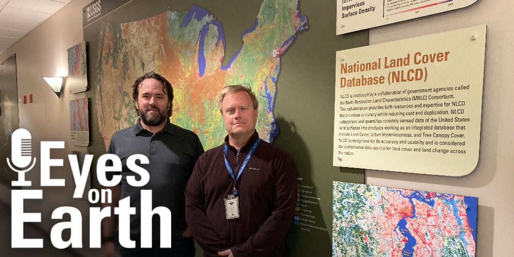 Two men stand by a wall display of a map and explanations, with a logo on top