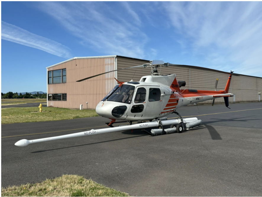 Red and white helicopter with long boom extending forward; helicopter is on the ground in front of building
