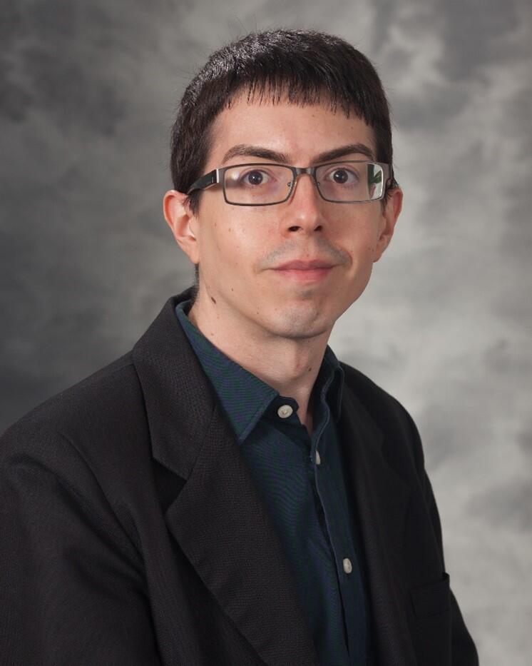 Portrait of man with dark hair and glasses wearing a suit.