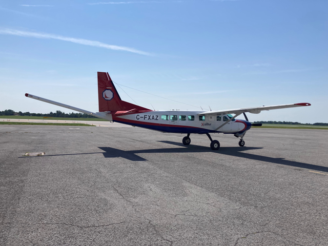 Image shows a small, single-engine fixed-wing aircraft with a stinger sticking out the back of its tail. The airplane sits on an airport tarmac under a blue sky.