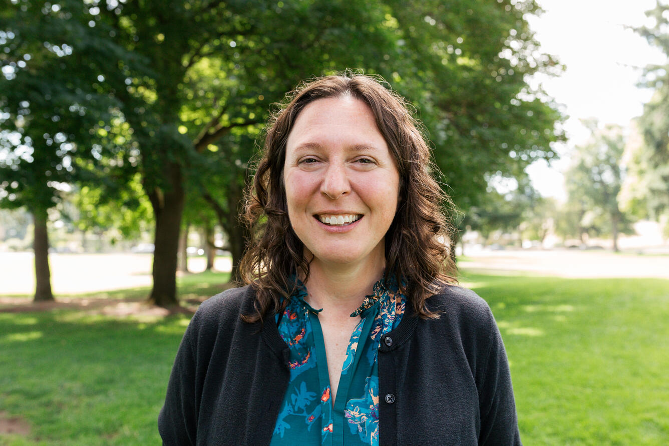 A woman with brown hair wearing a blue shirt and black cardigan smiles for her picture while in a park.