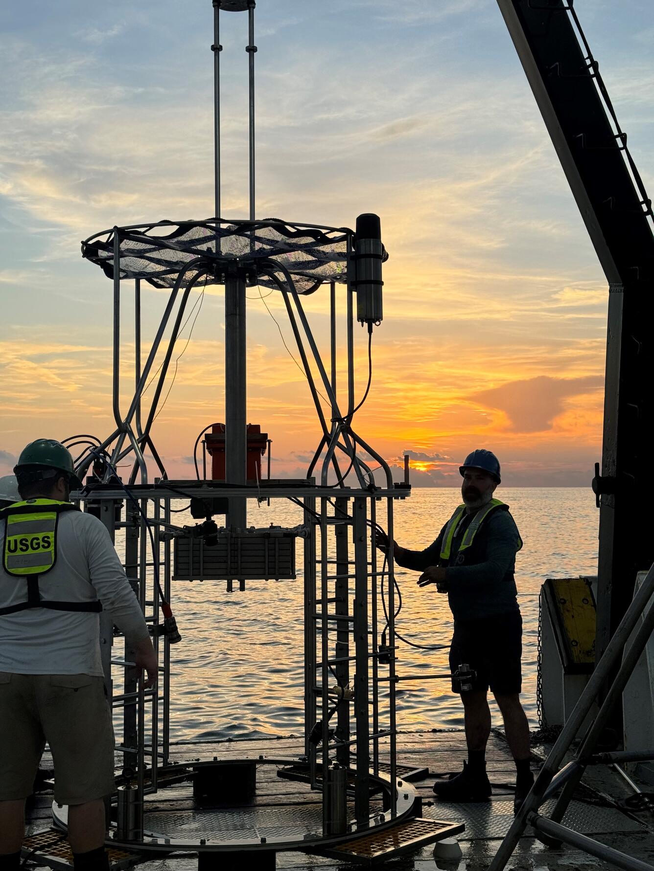two people working with a mega multicorer on the deck of a ship at sunset