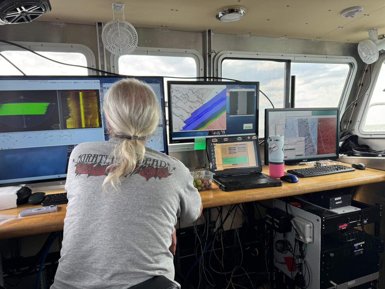 Person sitting at desk onboard a ship in front of multiple computers showing seafloor mapping data