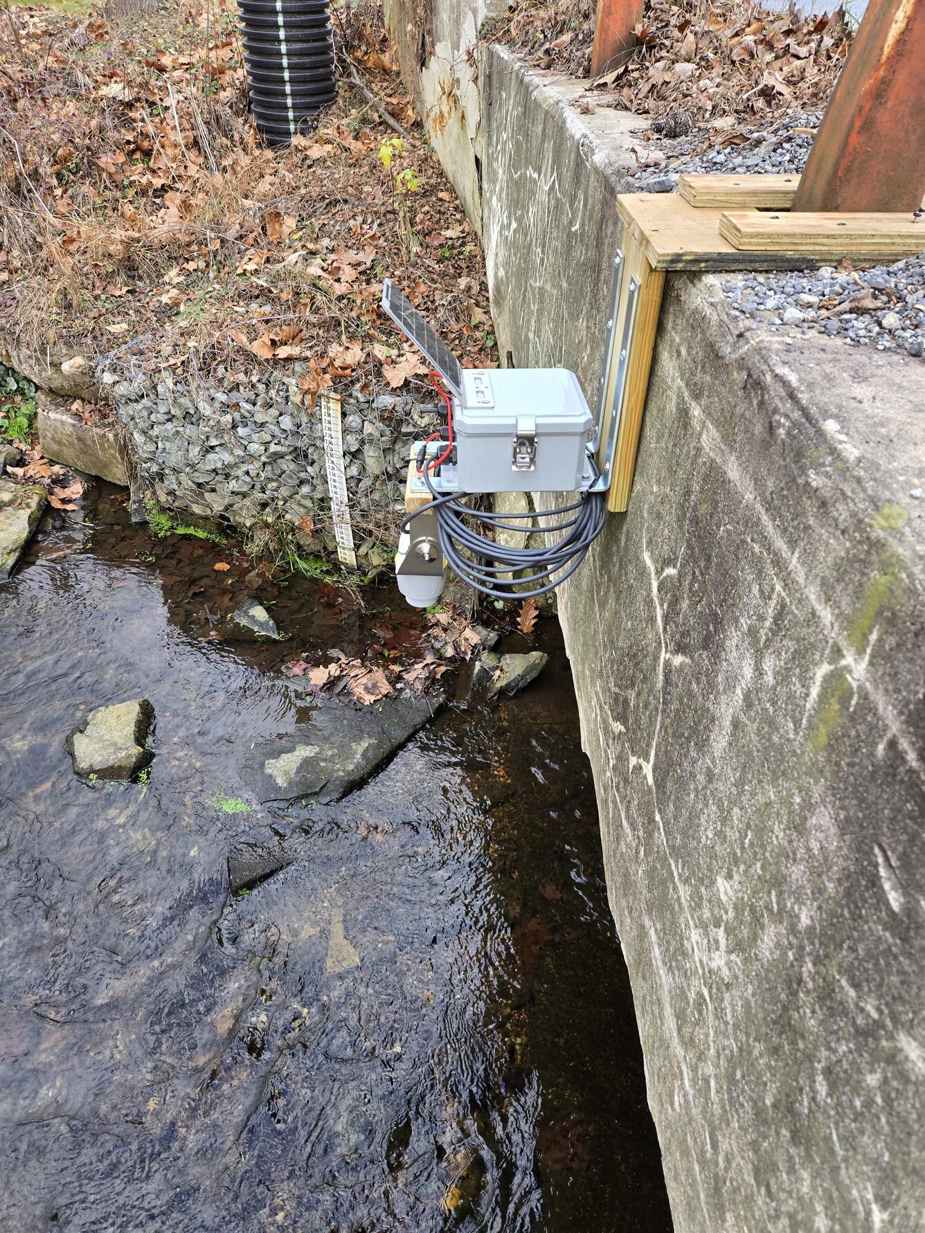 Gray box with attached solar panel attached to bridge with stream below
