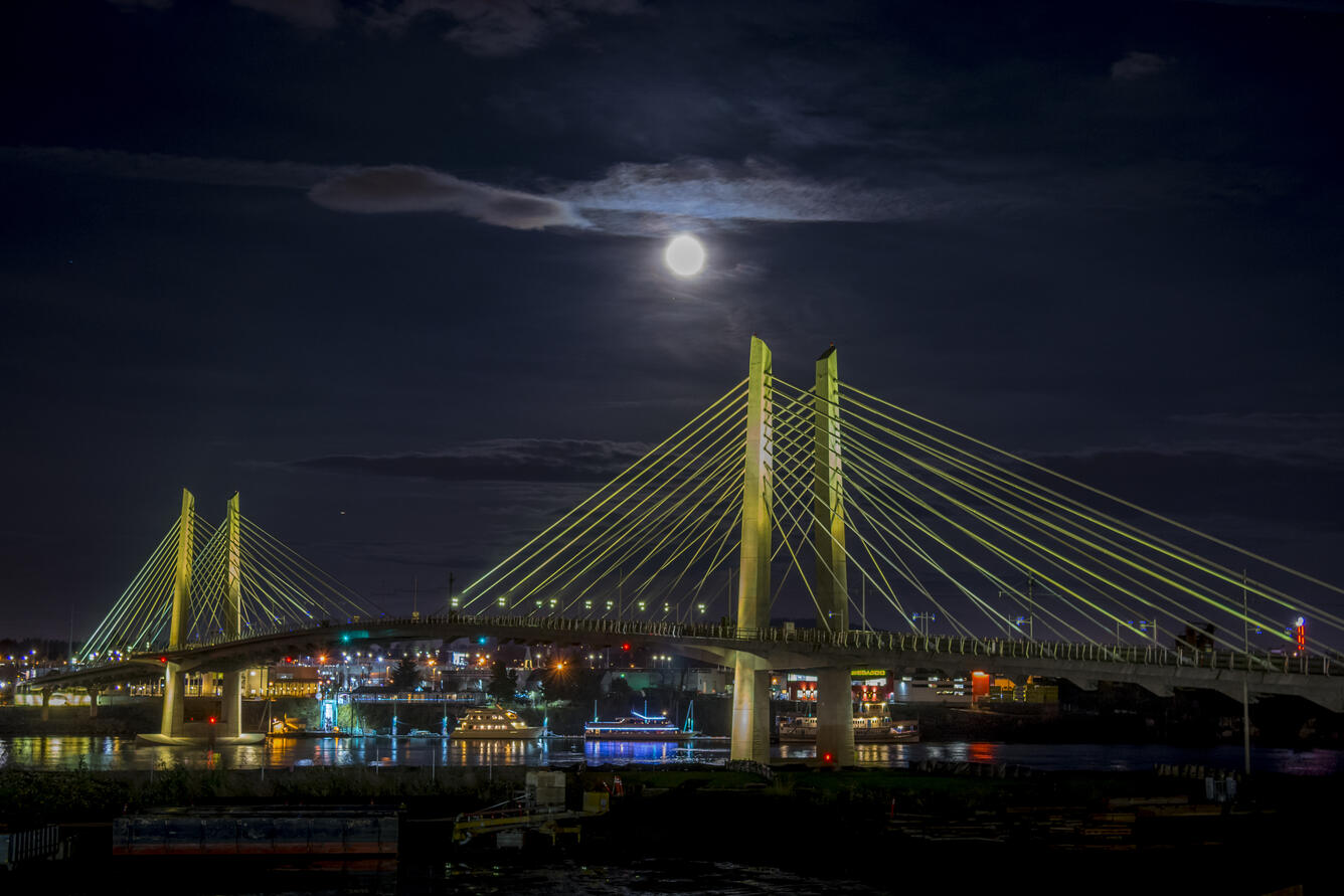 brightly lit colorful boats float below the bridge with its cables lit a pale yellowish green on a full moon night
