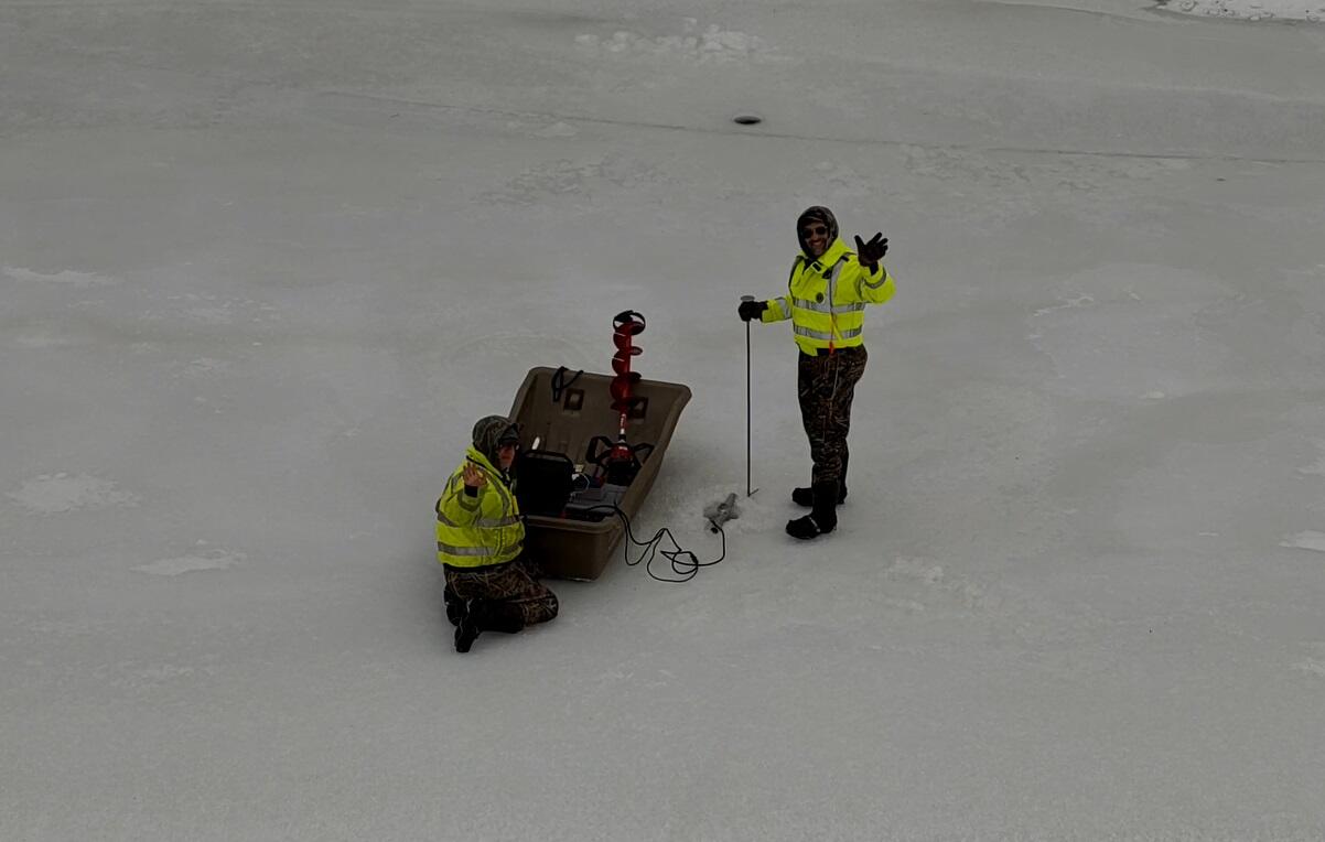 Two scientists next to an ice sled on the frozen Delaware River take a moment to wave at the camera