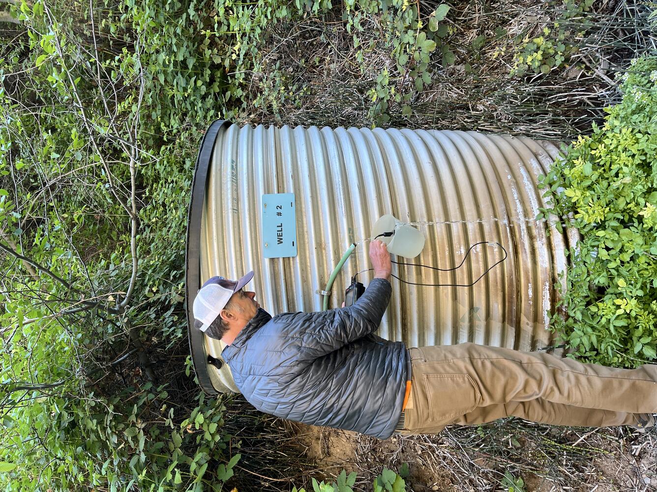 Tall man in a baseball cap uses a bucket to catch well water in to take measurements with a handheld sensor