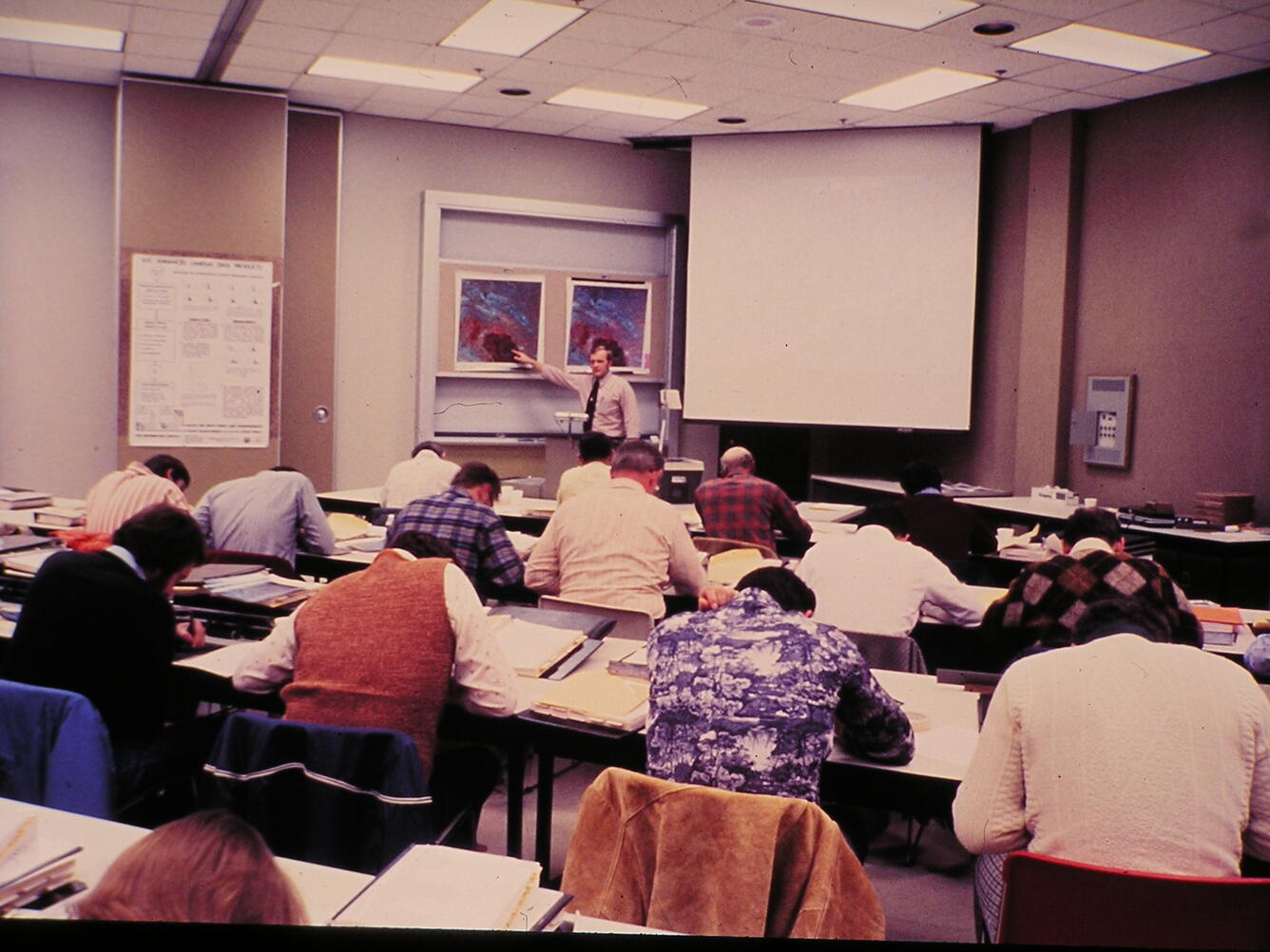 A man lectures at the front of a room of people seated and bending over tables.