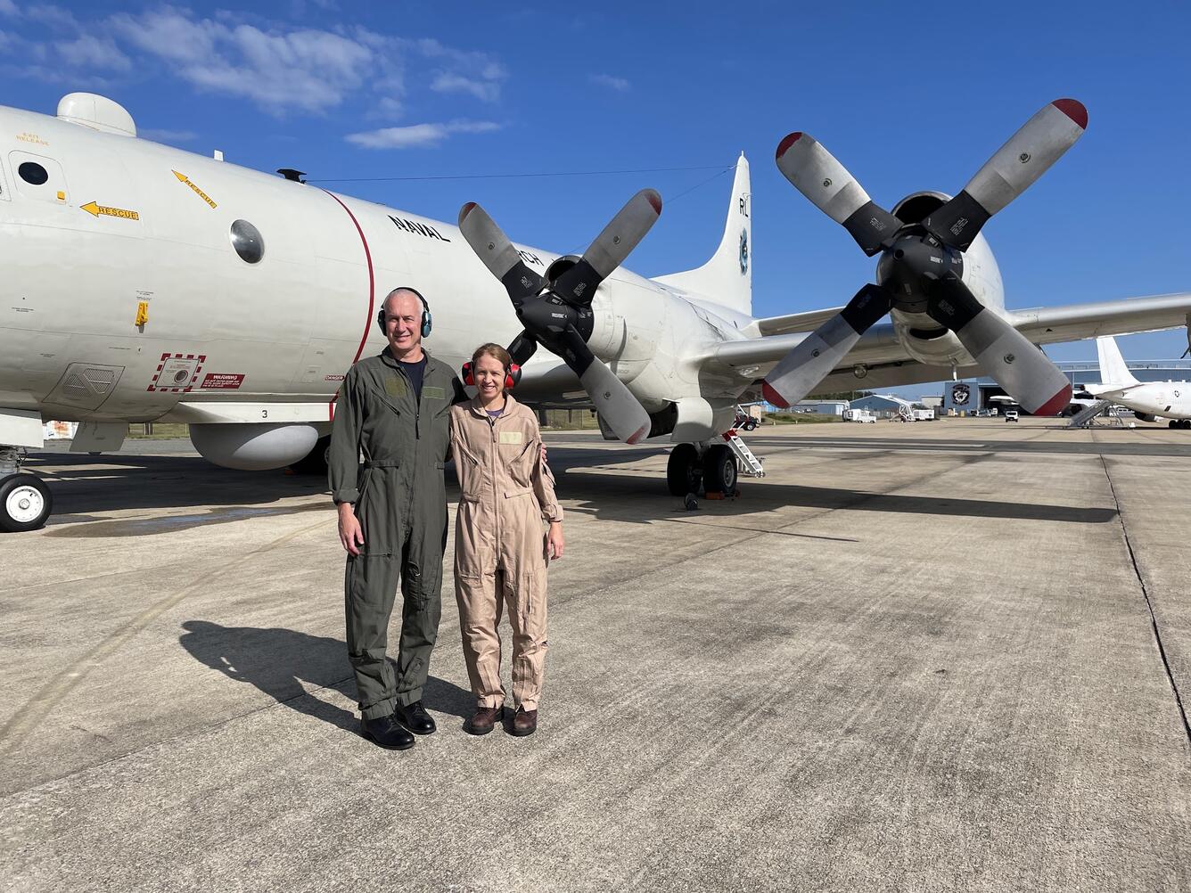 Two people in flight suits standing together smiling next to an aircraft on a runway