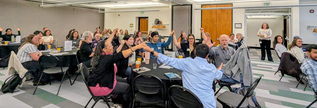 Groups of people at tables lift hands into the air to link up in unison
