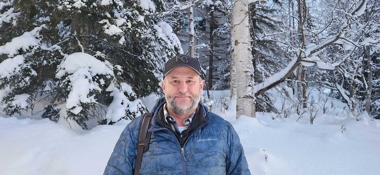 A man in a winter jacket and a baseball cap stands in the snowy woods for his picture.