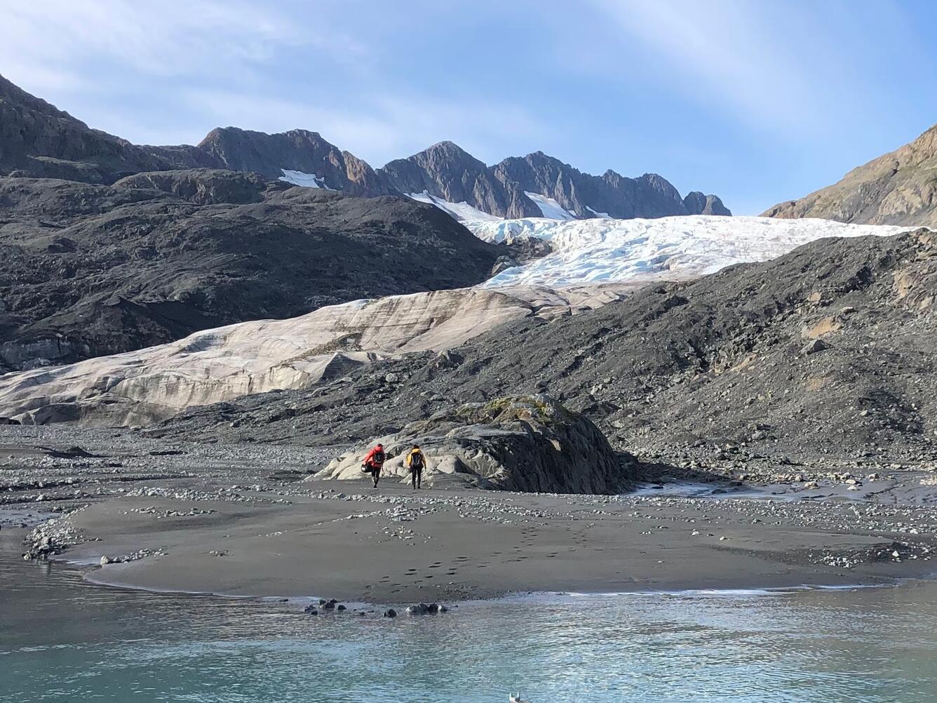People standing on shoreline near water with mountains and glacier in the background