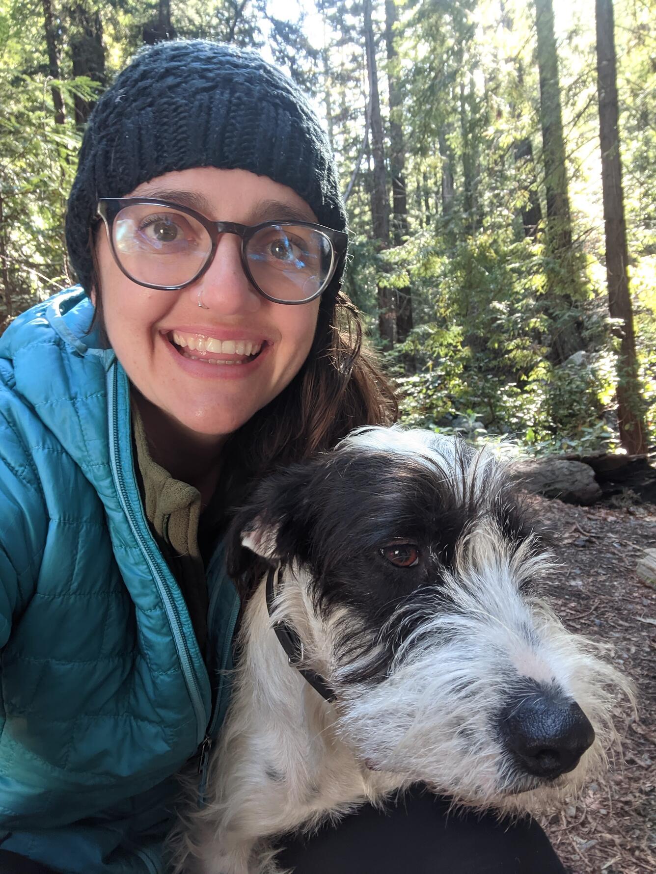 A Caucasian woman wearing a black beanie, glasses, and a light teal puffy jacket crouching with a white and black scruffy dog