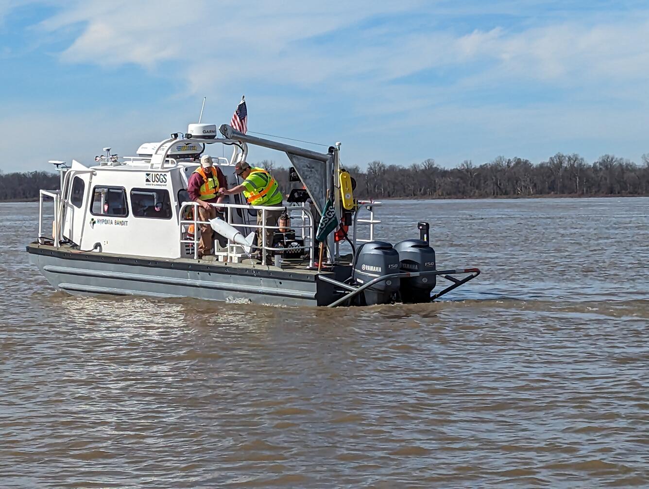 Two USGS scientists lower a water-quality and sediment sampler into a large river from the side of the boat