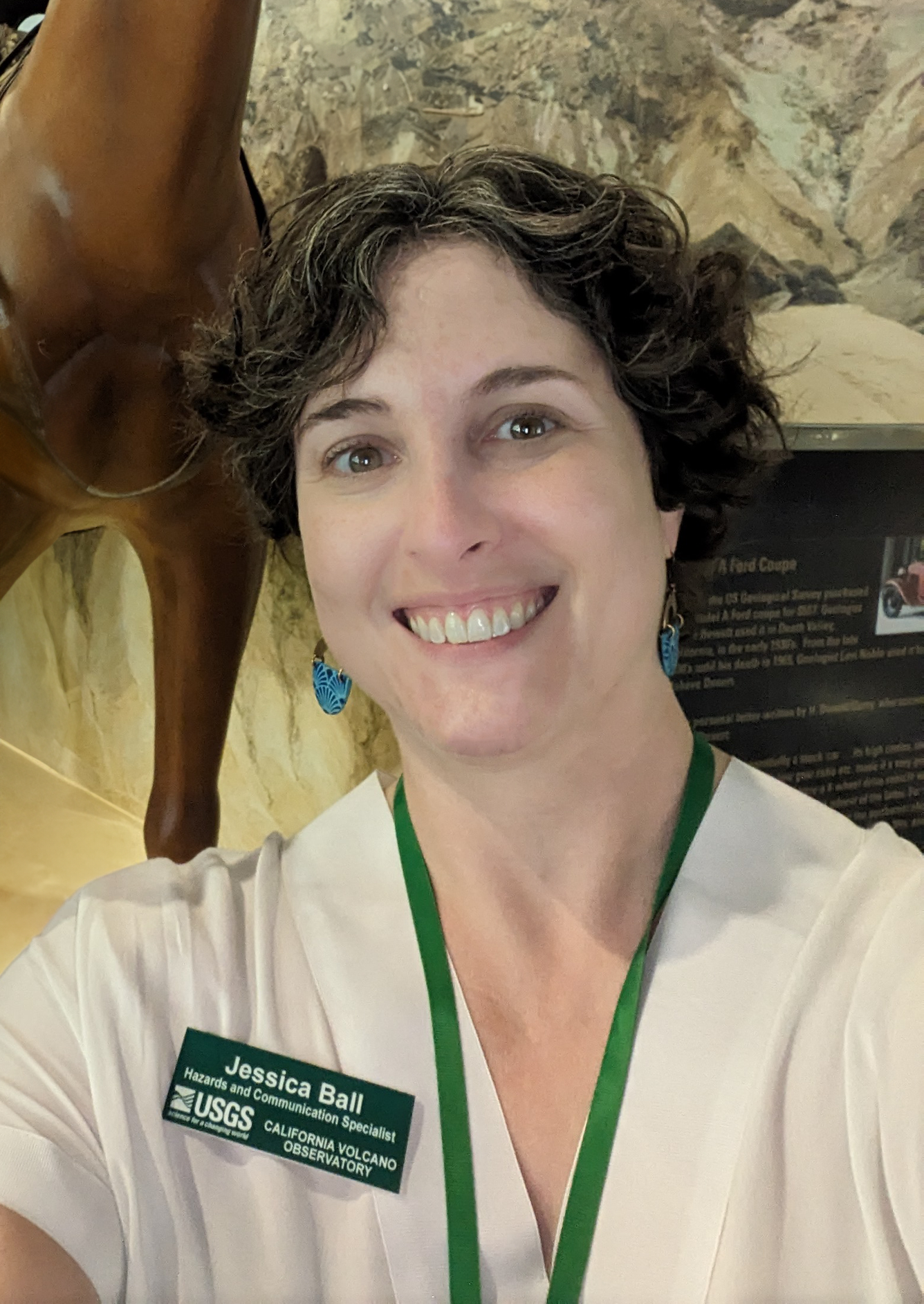 A woman with short, curly, salt-and-pepper hair smiles at the camera while standing in front of a museum exhibit featuring a fiberglass horse. She is wearing a white shirt with a green name badge and a lanyard.
