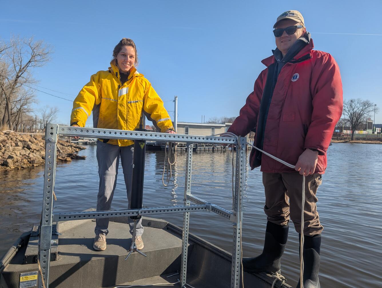 Two people hold up a metal frame while standing on a boat at the edge of the river