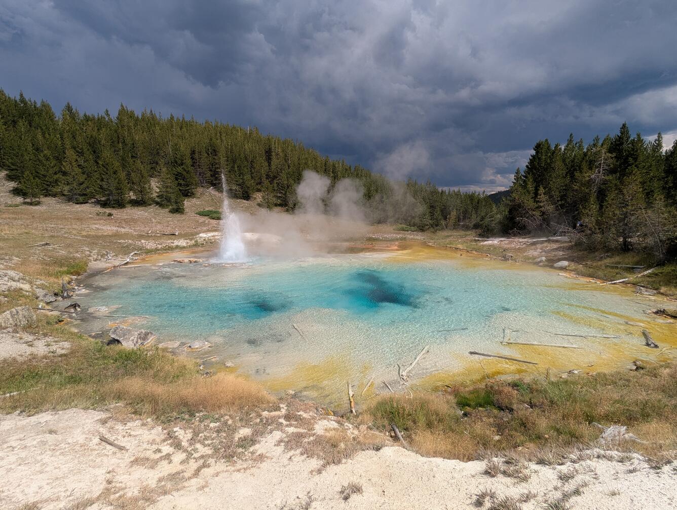 blue-water hot spring with a geyser to one side, forest in background, and storm clouds in the distance