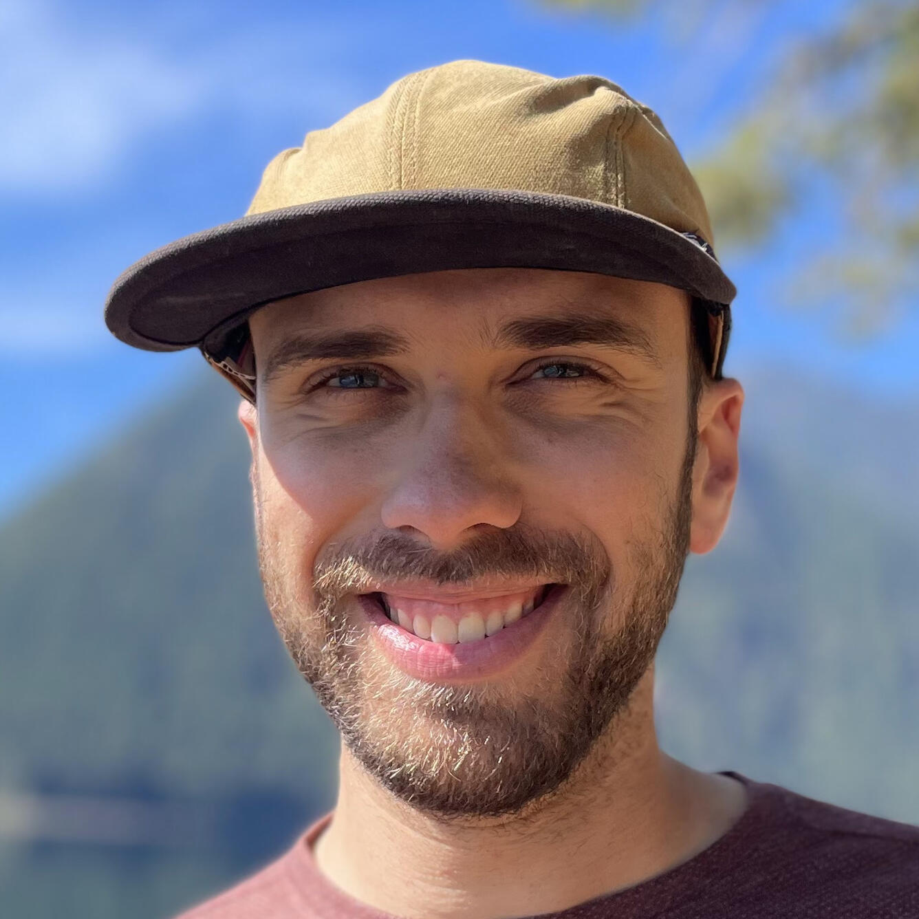 Photo of a young man wearing a fishing cap and smiling. He has blue eyes and brown hair and a beard. A mountain and lake are visible behind him.
