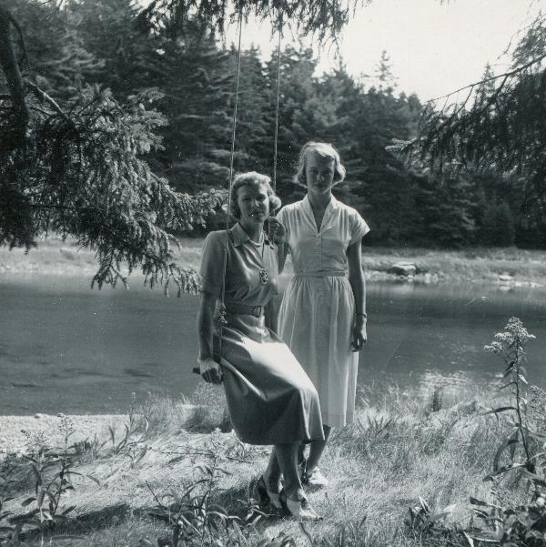 two women next to a lake, one is sitting in a swing hung off of a tree