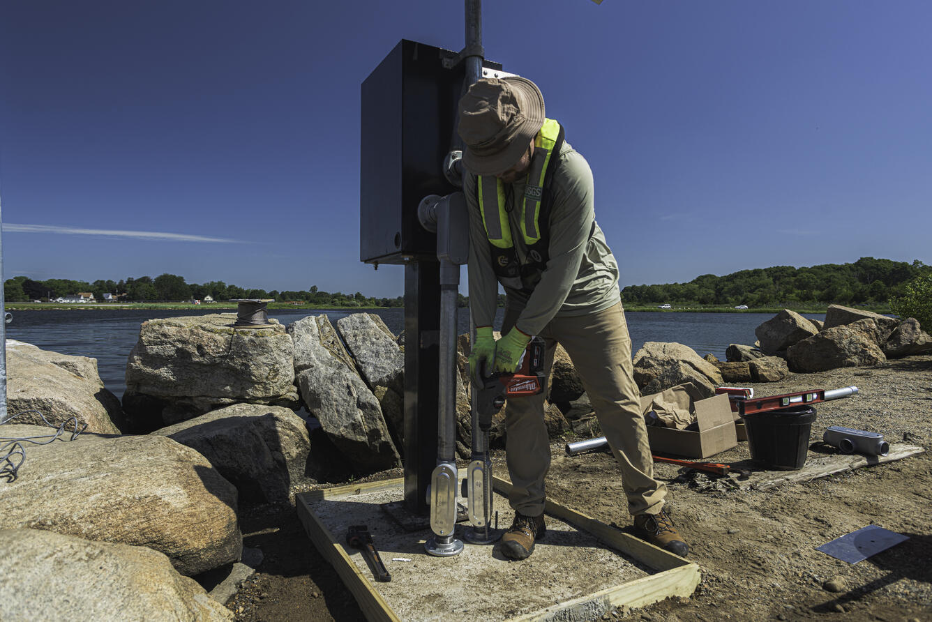 Person using drill to install anchor for gage monitor with river in the background.