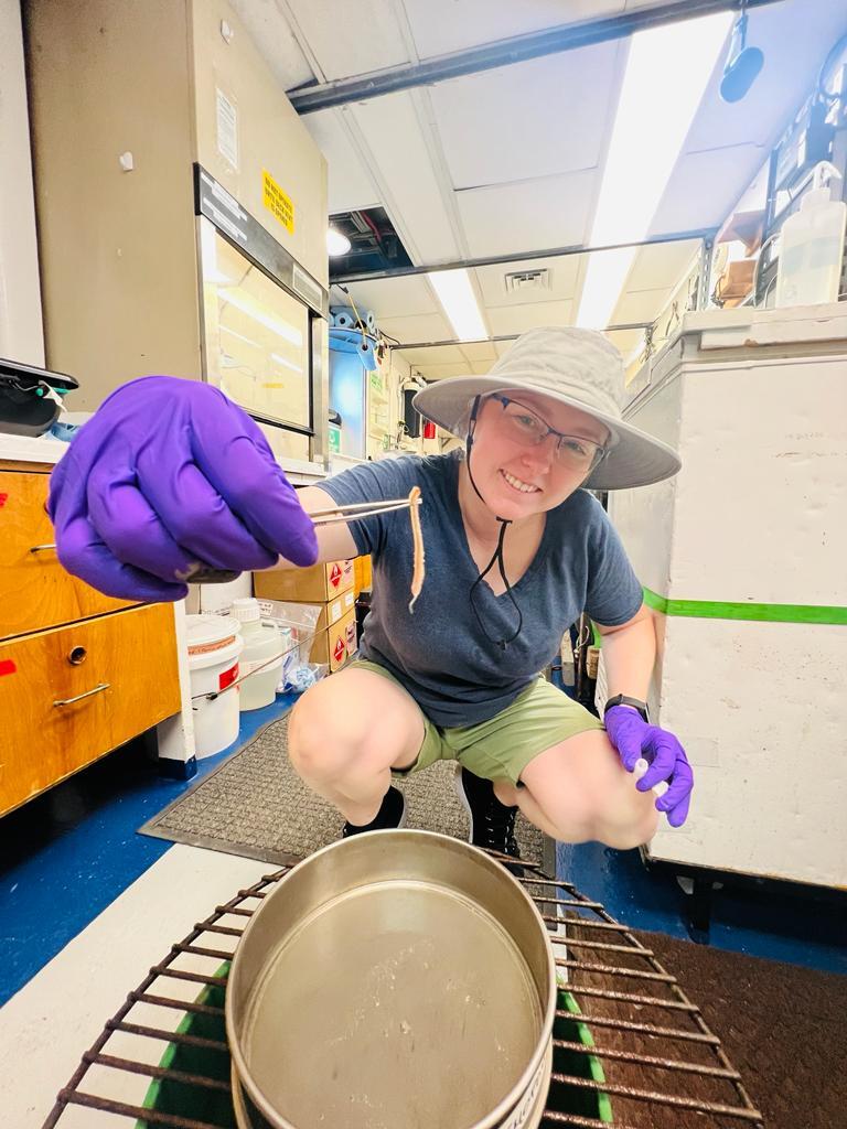 A person in a hat crouches down while holding a worm in their hand and smiles to the camera
