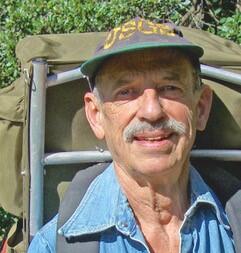 Headshot of a man with a moustache, wearing a USGS hat and an army-style backpacking pack