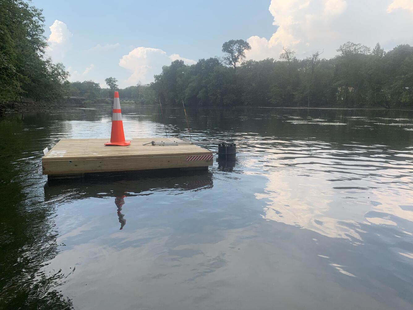 An orange traffic cone sits on top of a floating wooden platform supporting the sampler in the middle of the river