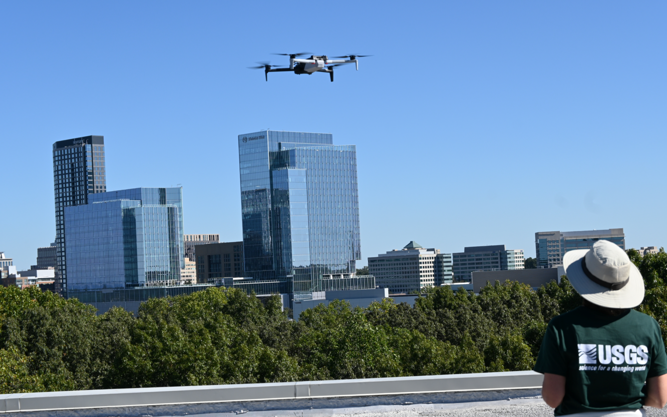 The drone operator, in a USGS shirt, looks at the black-and-white drone as it flies.