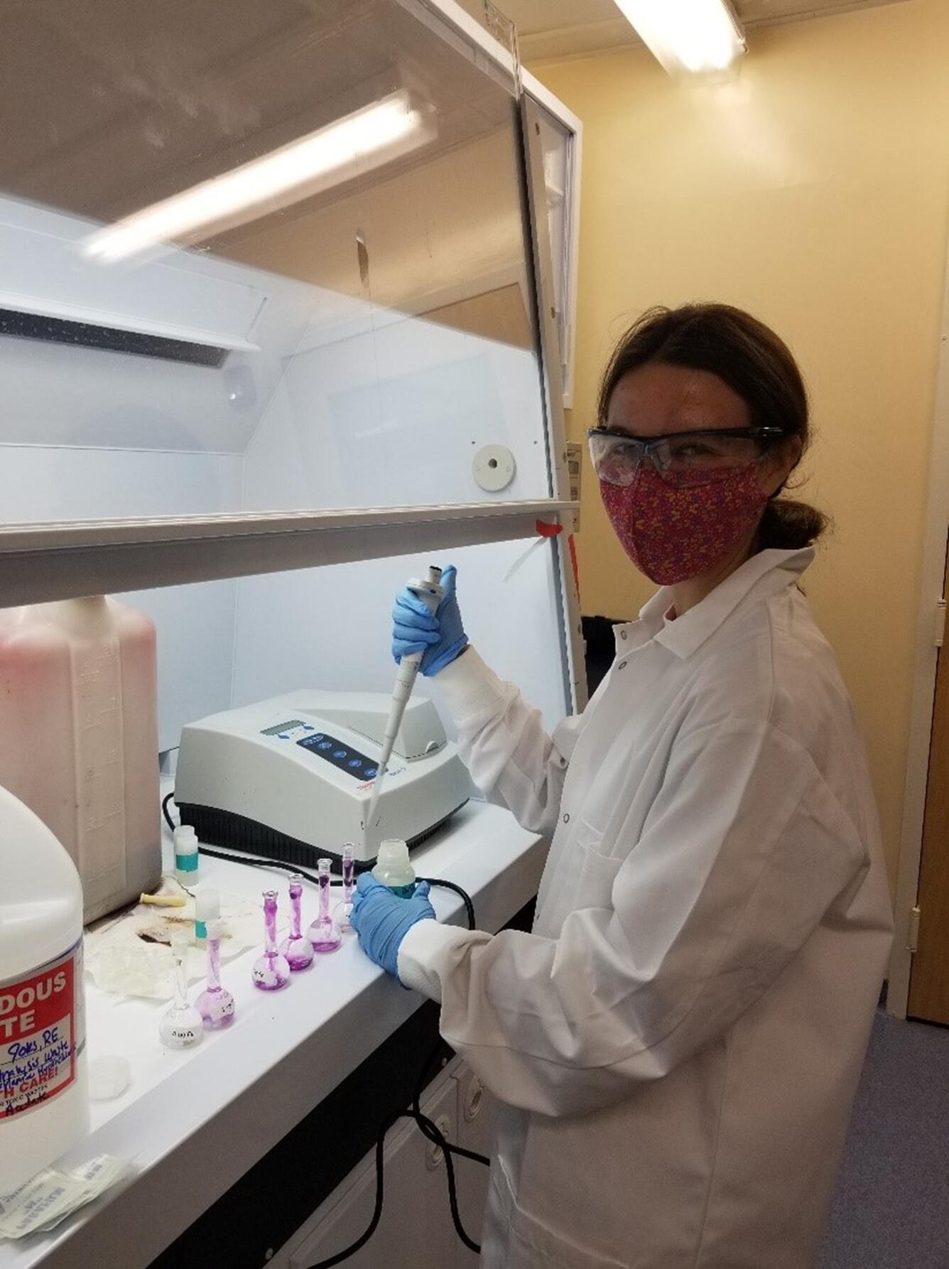 Person in lab coat working in lab, looking at camera with mask on