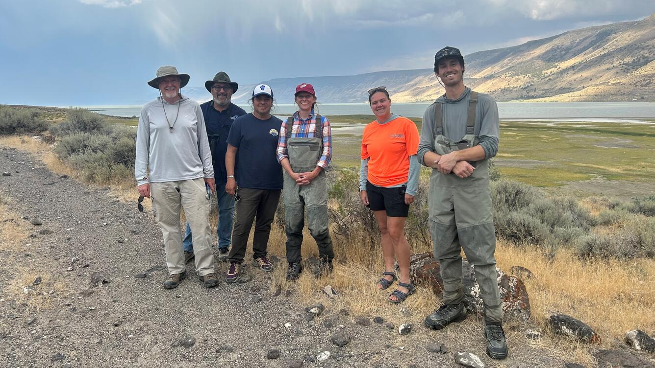 Five people in outdoor gear smile in front of a shallow lake on a sunny day