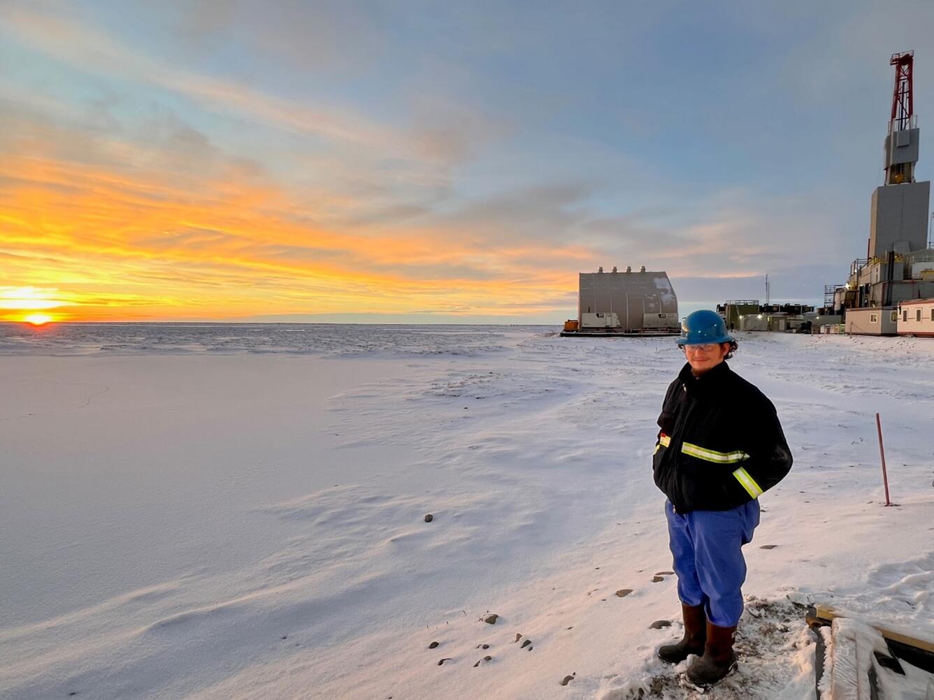 Person with hardhat on outside at sunset, snow on ground, some machinery in the background