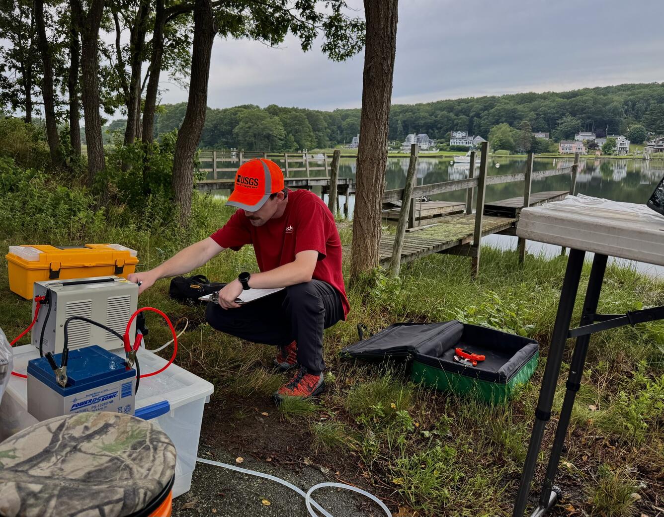 A man wearing a red shirt and orange hat crouches down in front of a peristaltic pump in front of a dock.