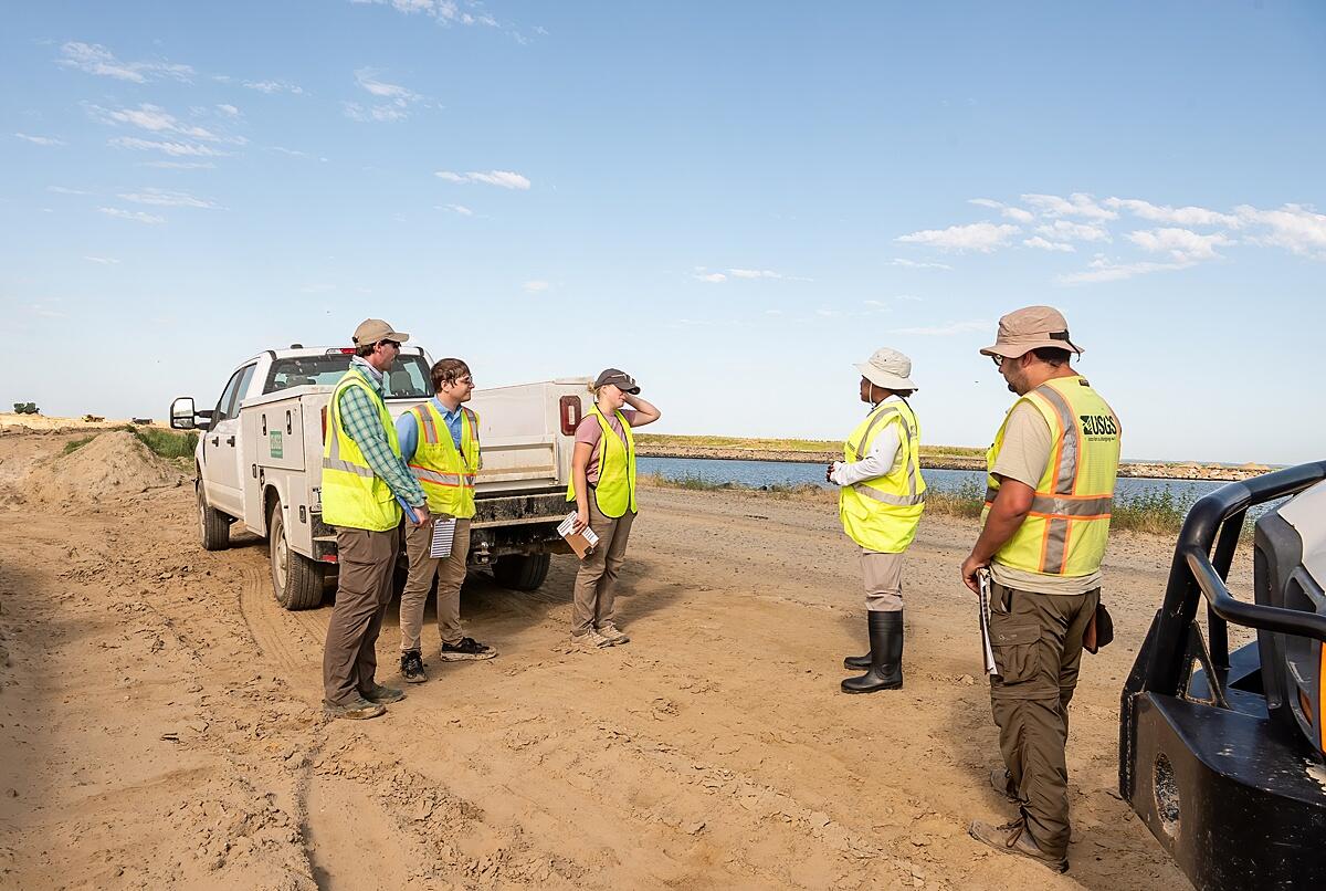 Five scientists in hats and bright green safety vests stand on the sand near two pick-up trucks.