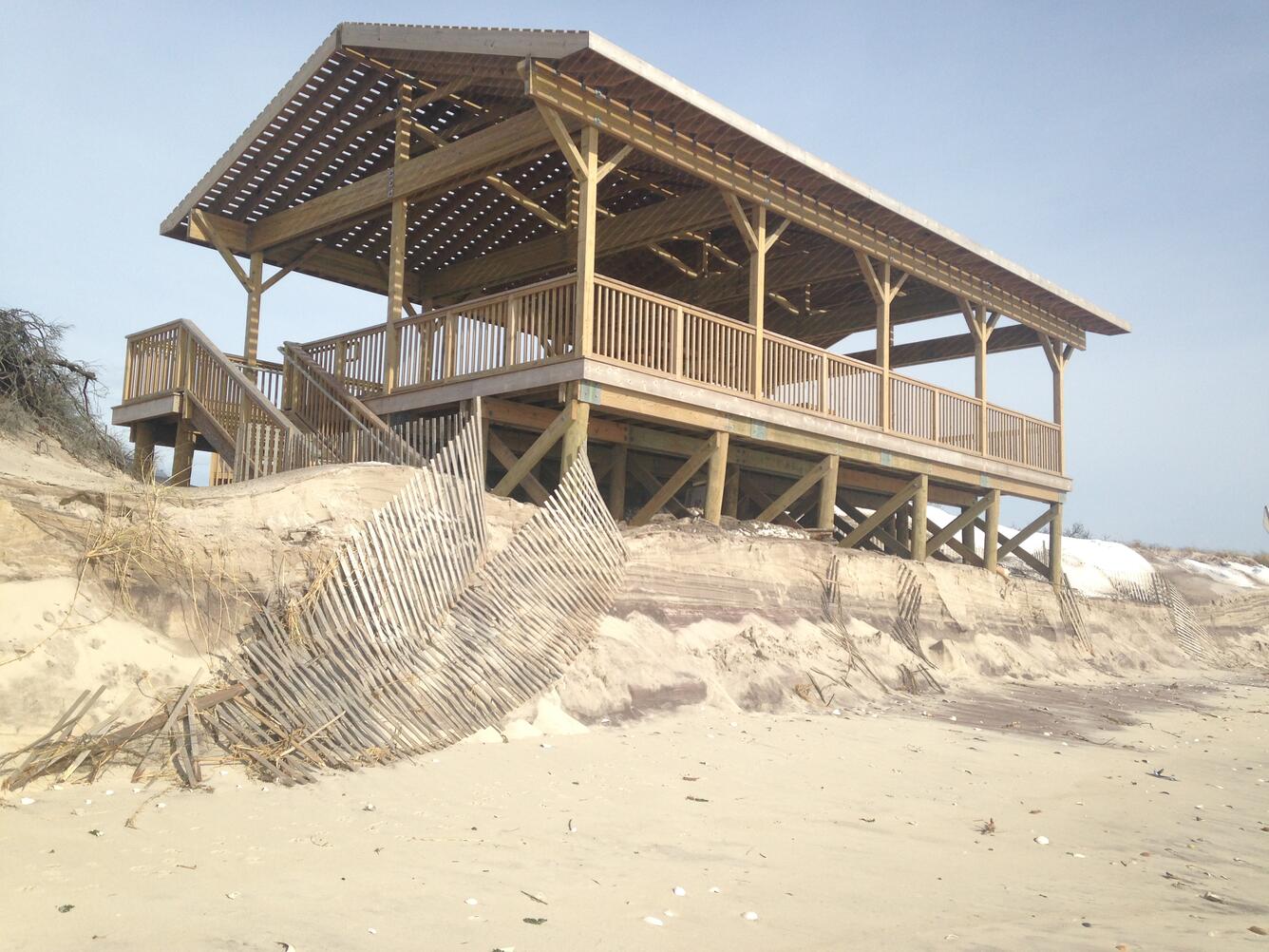 A pavilion on a beach at the edge of an eroded dune is partially covered by sand after a winter storm