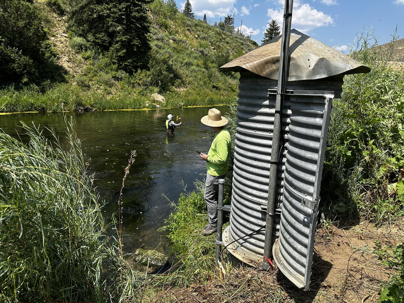 Corrugated galvanized steel gage house with tech next to it and person wading in river in background.