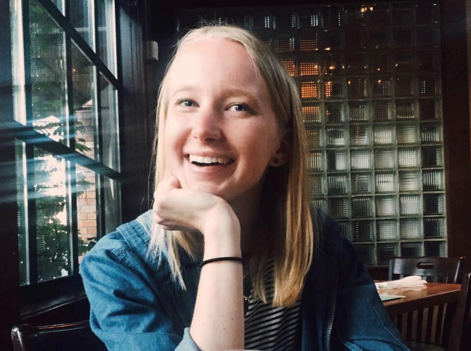 Woman in her late 20s, smiling at the camera, blonde hair and blue eyes, her hand is under her chin, and the background is windows with a blue tinge.