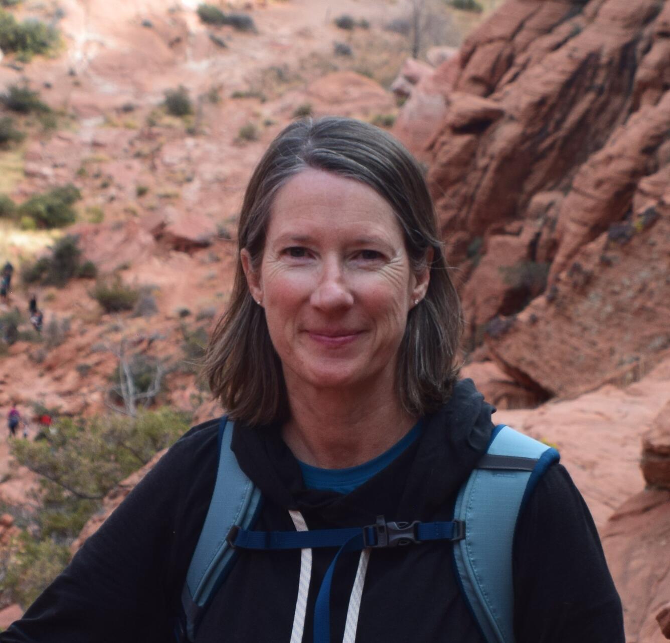 Woman wearing black shirt and blue backpack stands in front of red rocks