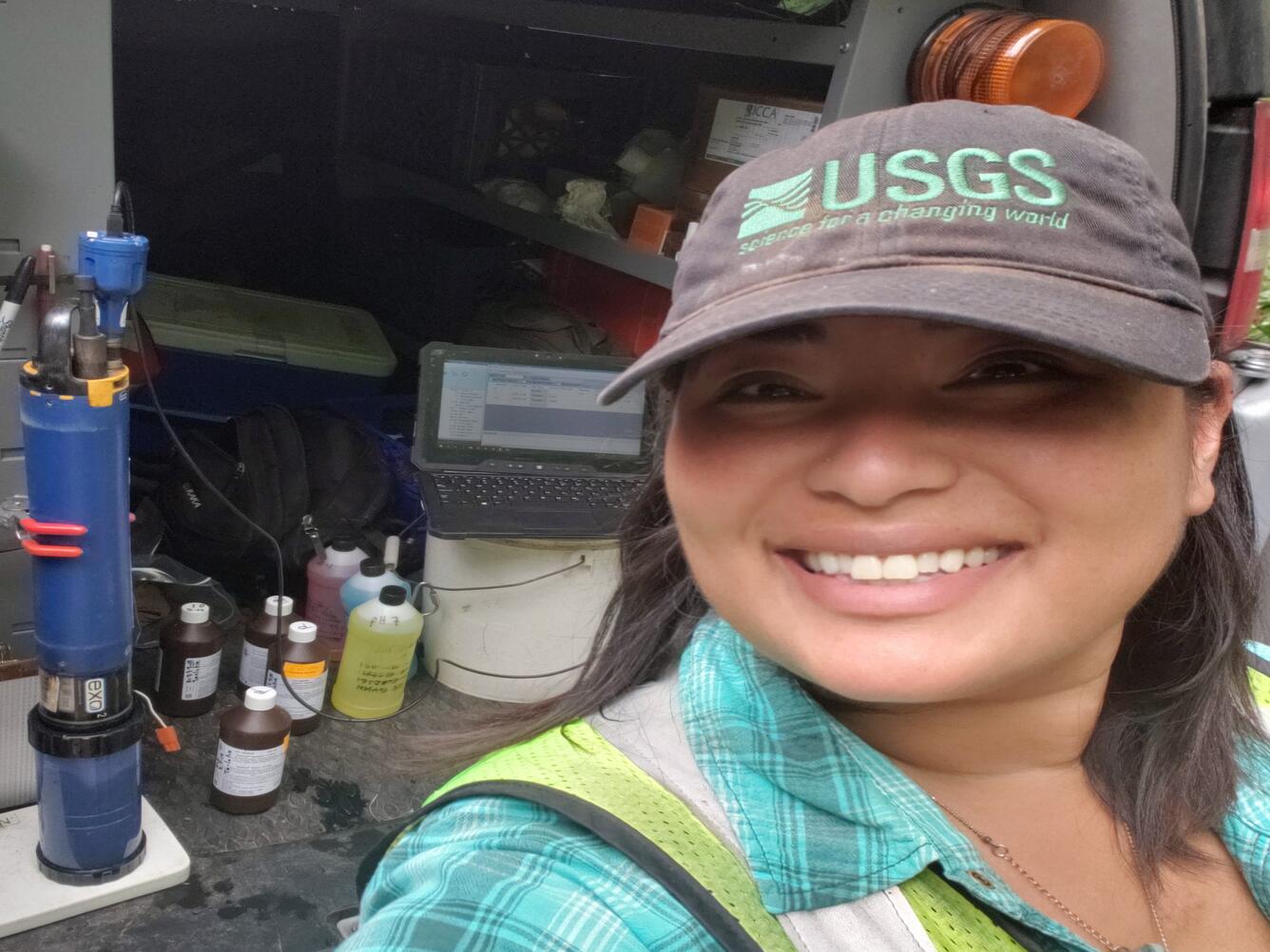 smiling female in a grey hat with green USGS logo wearing safety gear and teal plaid shirt stands by water quality sensors