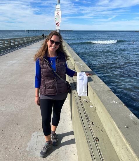 Katie stands on an lake pier with one arm resting on the rail. A smiling face against a backdrop of blue water.
