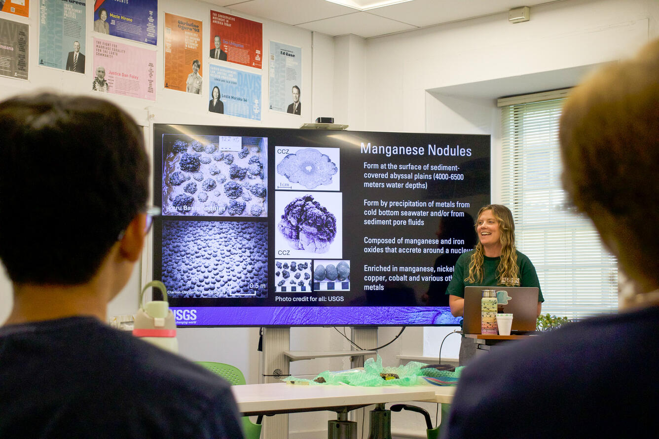 A women stands beside a large presentation screen in front of a classroom.
