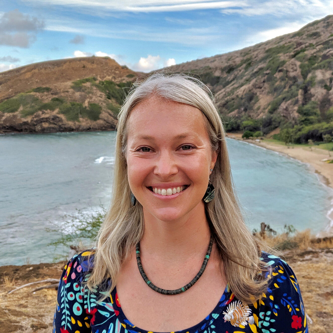 scientist with blonde hair, blue eyes, wearing a floral shirt, standing near water and mountains on a sunny day