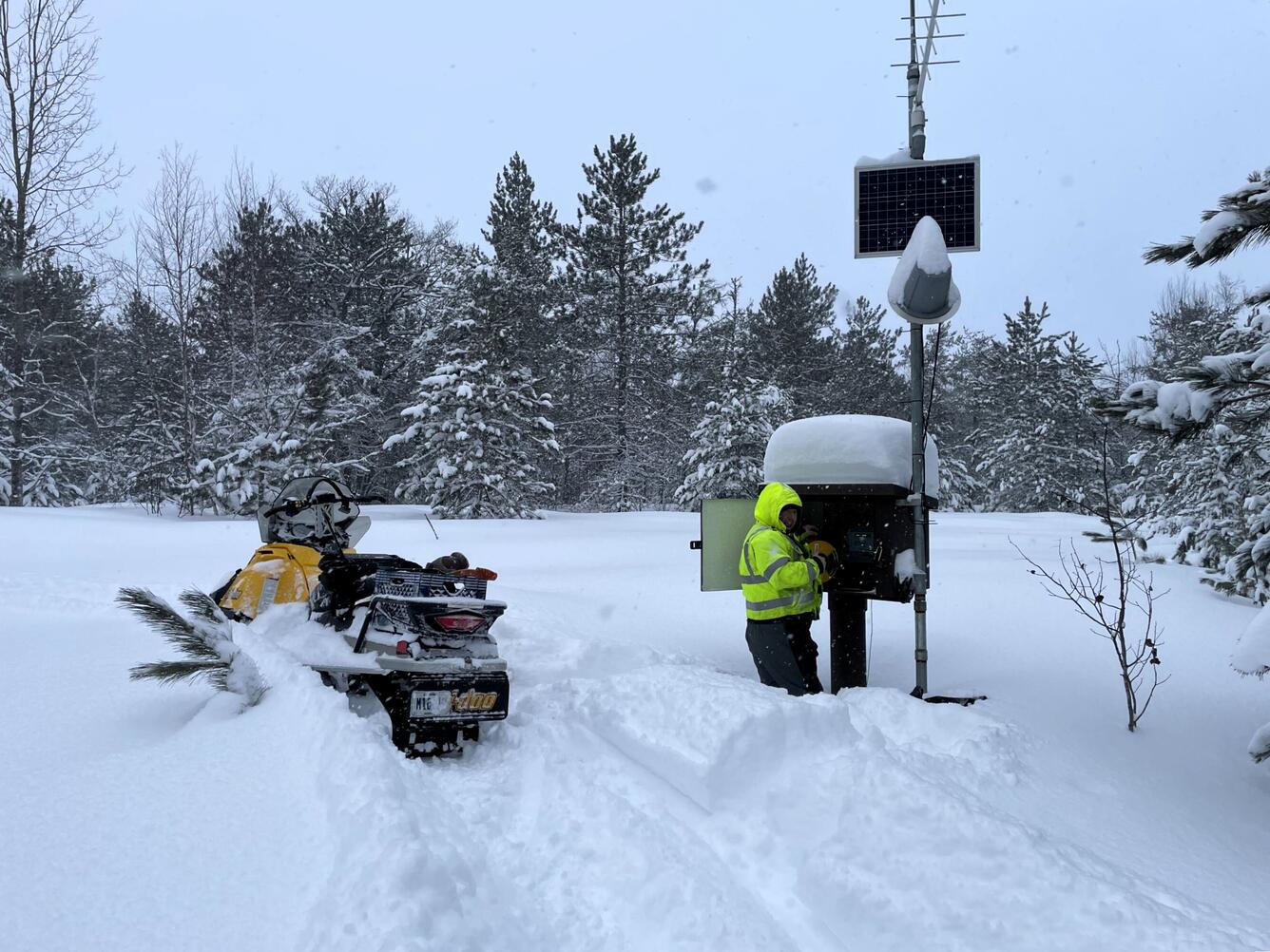 A Hydrologic Technician works on equipment inside a monitoring location housing in knee-deep snow next to a snowmobile