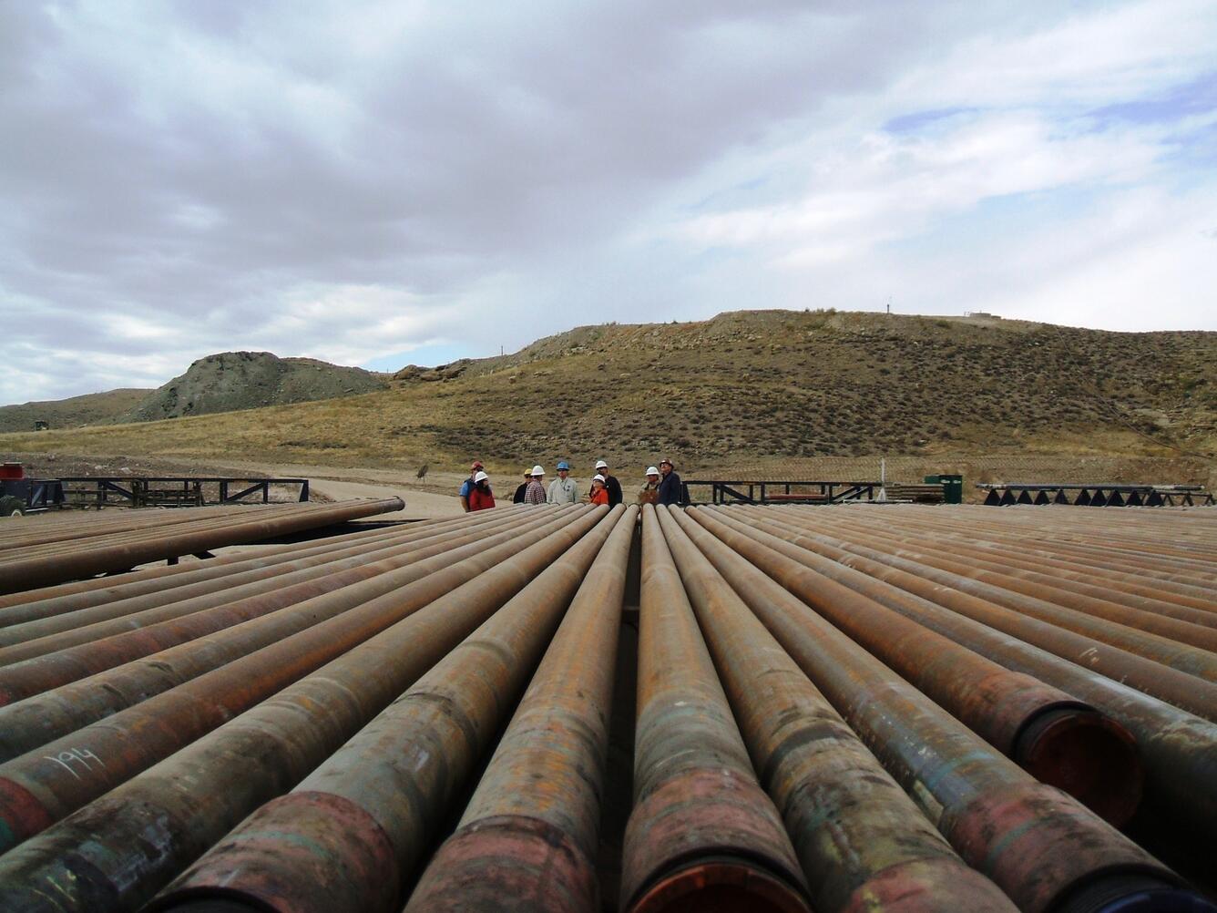 large metal pipes lying on a platform, with a few helmeted people and hills in the background