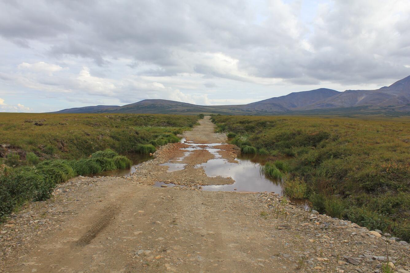 An uneven road with dips and puddles in a barren landscape with mountains in distance