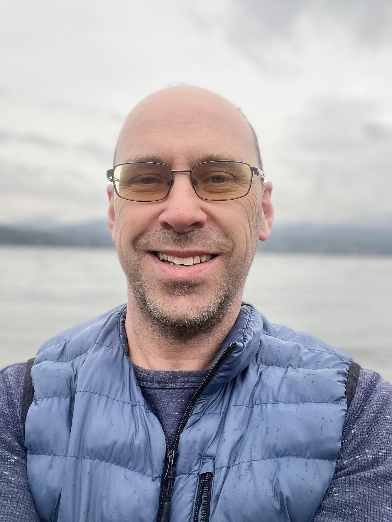 Man with blue vest and blue shirt and glasses smiling
