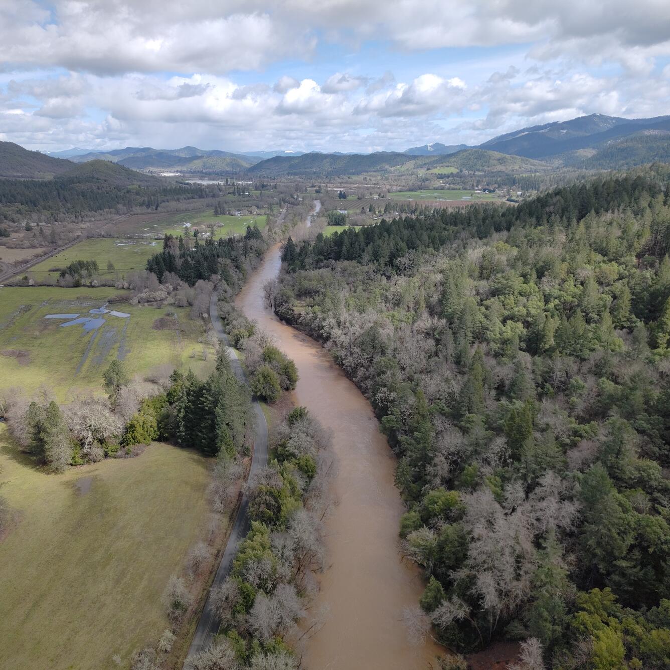 Brown river winds through forested valley with some open pasture on the left. Partly cloudy skies.