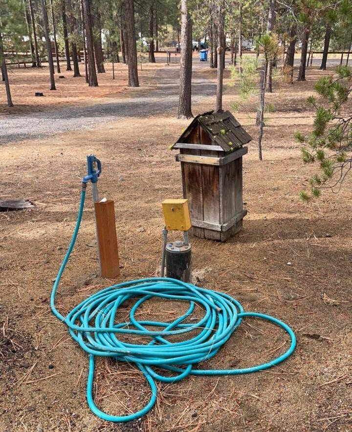 steel well casing with yellow electrical on top of well cap. In a wooded area with pine trees and light brown earth