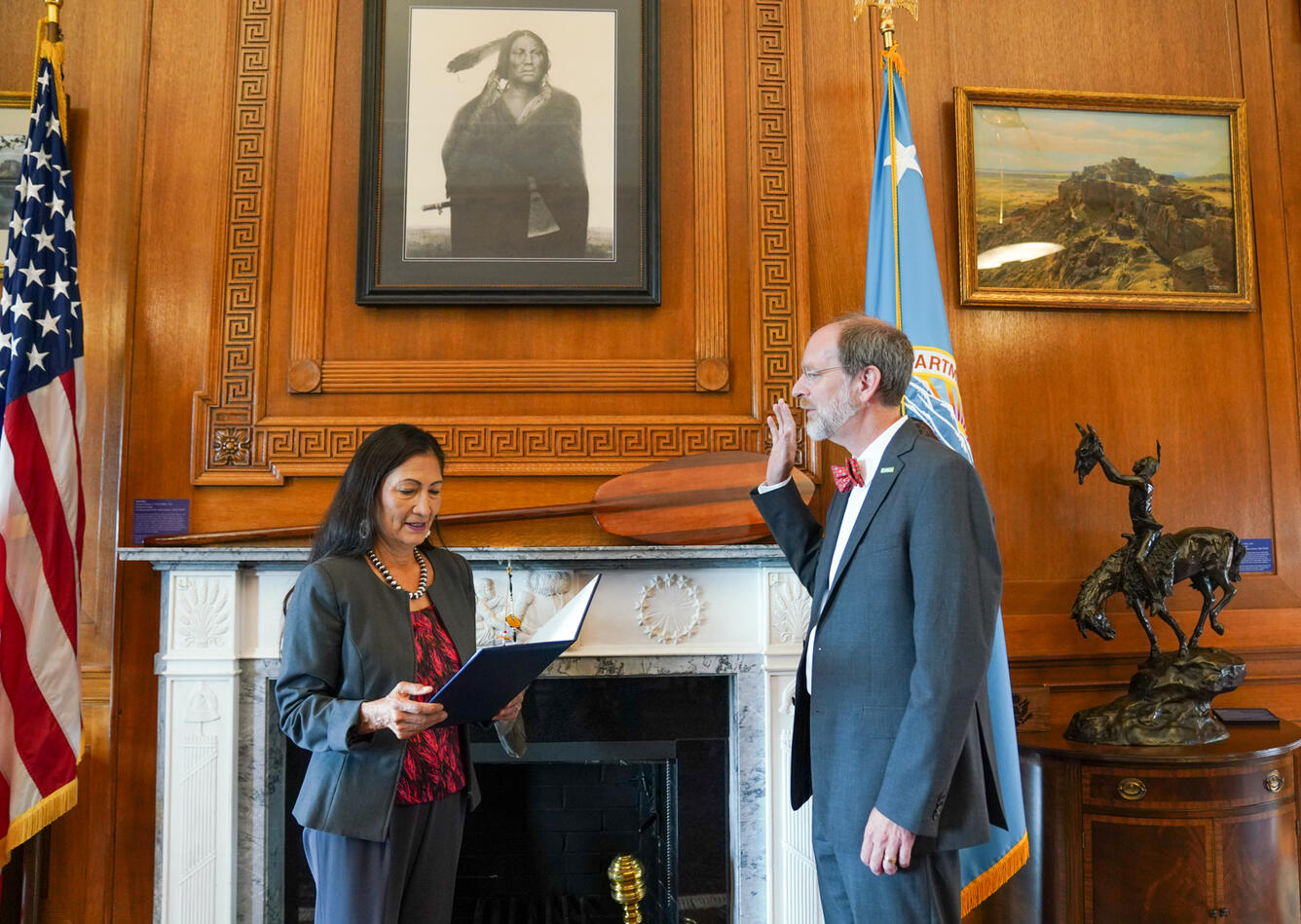 Image shows a man in a suit swearing an oath administered by a woman in a suit in a wood-paneled office